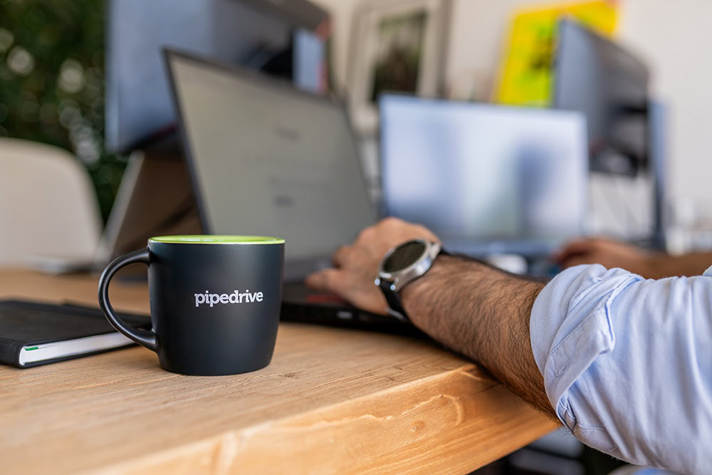 Close-up van een zwarte Pipedrive-mok op een houten bureau, naast een notitieboek, terwijl een persoon op een laptop werkt in een moderne kantooromgeving.