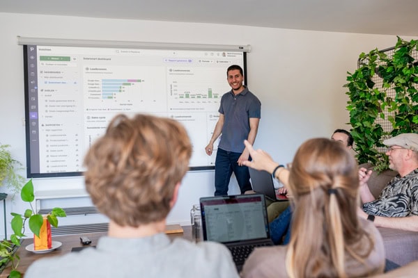 Man presenteert een dashboard met grafieken op een groot scherm aan een groep collega’s tijdens een teamoverleg in een informele vergaderruimte.