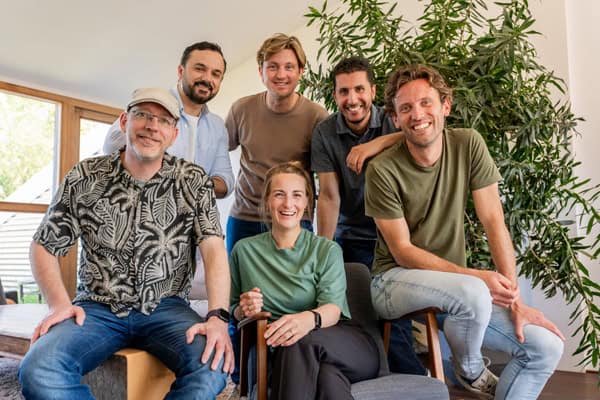 Groepsfoto van zes lachende collega’s in een informele kantoorruimte met planten en veel daglicht.